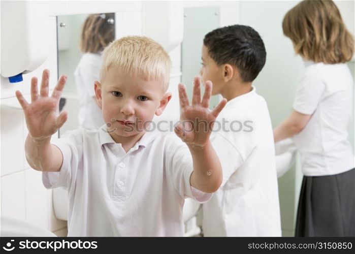 Students in bathroom at sinks washing hands with one holding up soapy hands (selective focus)