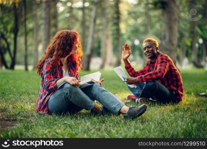 Students reading book on the grass in summer park. Male and female teenagers studying outdoors and having lunch. Students reading book on the grass in summer park