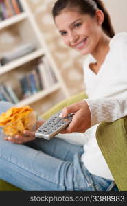 Students - Smiling female teenager watching television in living room with remote control and crisps