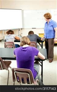 Students taking a standardized test as a teacher looks on to ensure no cheating.