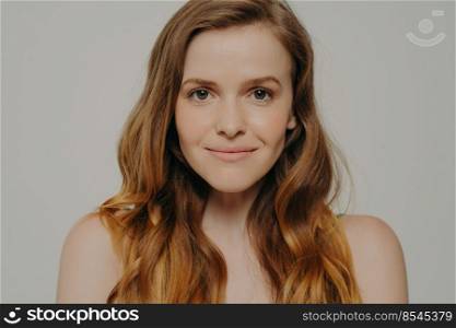 Studio headshot portrait of charming young woman with curly brown hair without makeup smiling at camera isolated over grey background, beauty shot of gorgeous caucasian female with healthy skin. Beauty shot of gorgeous caucasian female with healthy skin isolated on grey background
