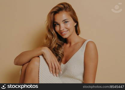 Studio photo of beautifully tanned slender charming young lady sitting on chair with legs up in dress with open shoulders, slightly tilted her head looking at camera with beaming smile. Beautifully tanned charming young lady sitting on chair with legs up, isolated on beige background