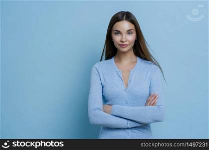 Studio photo of pretty female model stands with crossed arms, has long hair, being confident and self assured, wears blue sweater, looks at camera, poses indoor, blank space for your promotional text