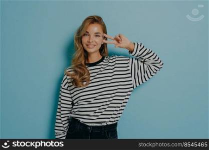 Studio portrait of attractive charming smiling girl in casual striped longsleeve gesturing v-sign or peace symbol with two fingers close to eye, isolated on light blue wall background. Youth people. Portrait of attractive charming smiling woman in casual clothes gesturing v-sign isolated on blue