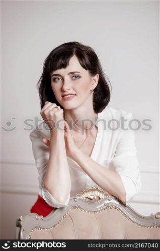 Studio portrait of attractive young woman with dark hair in white shirt and red pants leans on a classic chair on plain white background.Bossy female, job interview concept,confident independent lady. Studio portrait of attractive young woman with dark hair in white shirt and red pants leans on a classic chair on plain white background.Bossy female, job interview concept,confident independent lady.