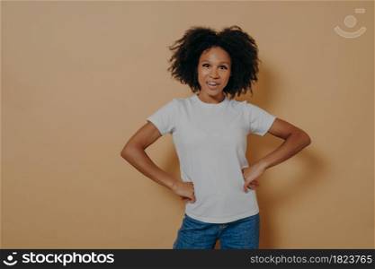 Studio portrait of beautiful confident african american female wearing casual stylish clothes keeping hands on hips and looking at camera with confidence, isolated on sand color background. Beautiful confident african american female wearing casual stylish clothes keeping hands on hips