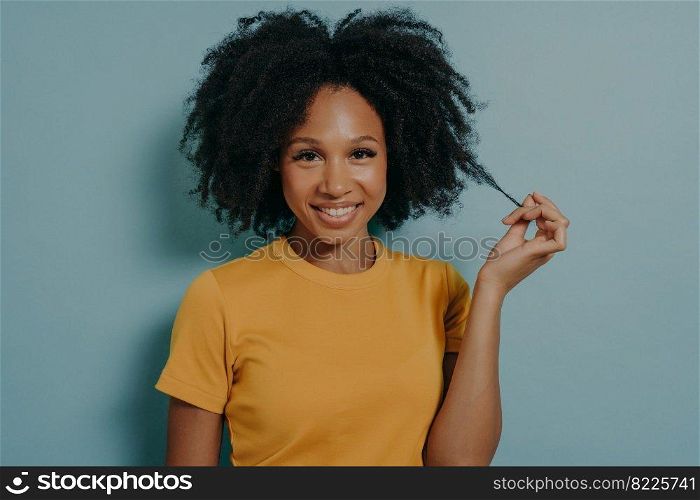 Studio portrait of beautiful young dark skinned woman with shaggy hairstyle smiling cheerfully, showing white teeth at camera, while playing with lock of her hair, feeling happy and excited. Studio portrait of beautiful young dark skinned woman with shaggy hairstyle smiling cheerfully