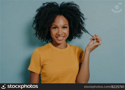 Studio portrait of beautiful young dark skinned woman with shaggy hairstyle smiling cheerfully, showing white teeth at camera, while playing with lock of her hair, feeling happy and excited. Studio portrait of beautiful young dark skinned woman with shaggy hairstyle smiling cheerfully