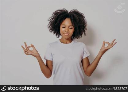 Studio portrait of calm and peaceful young mixed race woman keeping hands in mudra gesture and eyes closed, meditating while standing isolated over white wall. Meditation concept. Calm and peaceful young mixed race woman keeping hands in mudra gesture and eyes closed