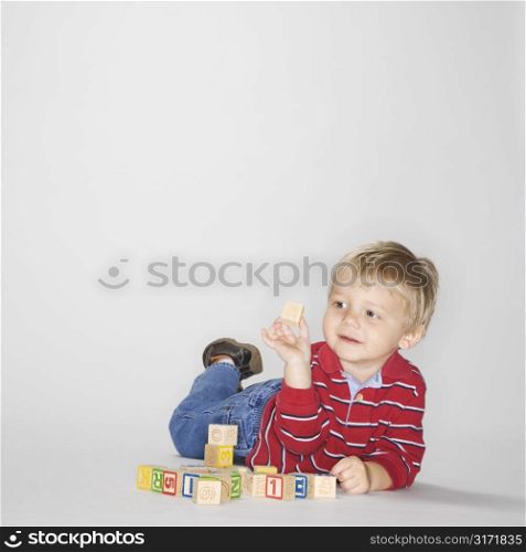 Studio portrait of Caucasian boy lying on stomach propped up on elbows playing with toy blocks.