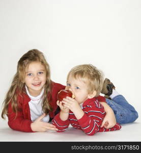 Studio portrait of Caucasian girl with arm around boy eating apple against white background.