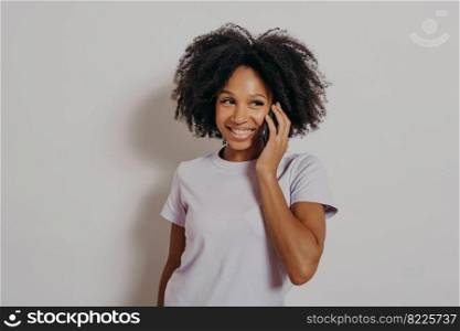 Studio portrait of cheerful dark skinned woman enjoying conversation on mobile phone, holding smartphone at smiling, hearing good news from friend while standing isolated over white background. Studio portrait of cheerful dark skinned woman enjoying conversation on mobile phone