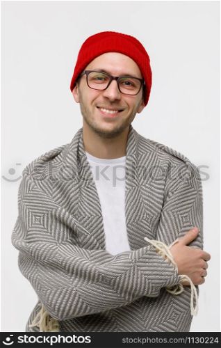 Studio portrait of cheerful stylish hipster guy with light bristles on the face smiling at camera, wearing glasses, red beanie and wrapped in plaid, arms crossed. Isolated on white background.