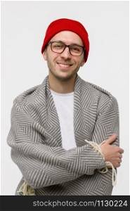 Studio portrait of cheerful stylish hipster guy with light bristles on the face smiling at camera, wearing glasses, red beanie and wrapped in plaid, arms crossed. Isolated on white background.