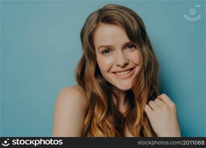 Studio portrait of cute teen girl with wavy hair smiling and looking at camera, headshot of romantic female posing against blue wall, expressing positive emotions and happiness, being in good mood. Studio portrait of cute teen girl with wavy hair smiling and looking at camera, isolated on blue
