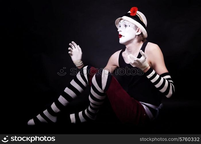Studio portrait of Dramatic funny mime in white hat with red flower and striped gloves sitting on floor on black background