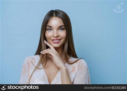 Studio portrait of gorgeous brunette lady touches chin, shows her beauty, cares about skin, feels tenderness, wears fashionable blouse, isolated over blue background being in relaxed and romantic mood
