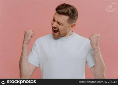 Studio portrait of happy overjoyed man in white blank tshirt enjoying victory or success, excited guy clenching fists and making yes gesture while standing isolated over pastel pink background. Young excited man in white blank tshirt celebrating success