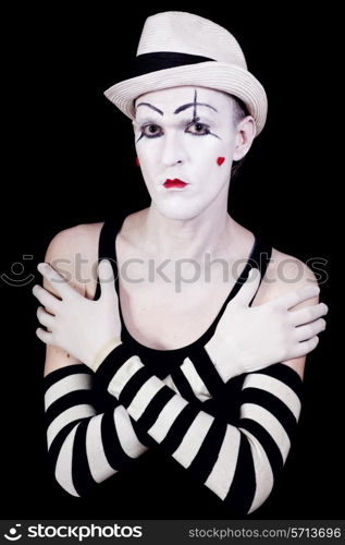 Studio portrait of serious theatrical clown in white hat and striped gloves with red hearts on her face isolated on black background