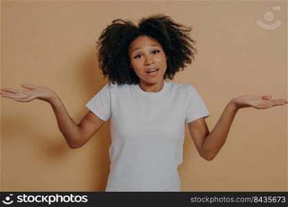 Studio portrait of unsure dark skinned woman shrugging shoulders and spreading arms sideways, can not give answer on question or having doubts while standing isolated over beige background. Unsure african woman woman shrugging shoulders and spreading arms sideways, isolated over beige wall