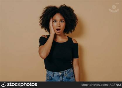 Studio portrait of young shocked african female with unexpected facial expression because of hearing bad news, mixed race woman keeping jaw dropped, wears casual outfit, posing against beige wall. Studio portrait of young shocked african female with unexpected facial expression