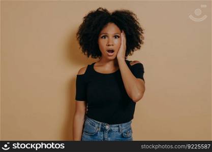 Studio portrait of young shocked african female with unexpected facial expression because of hearing bad news, mixed race woman keeping jaw dropped, wears casual outfit, posing against beige wall. Studio portrait of young shocked african female with unexpected facial expression