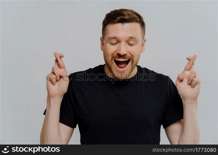 Studio portrait young man crossing fingers and making wish, excited guy hoping for win or begging for help while standing against grey studio background. Luck and superstition concept. Handsome young excited man in black t shirt crossing fingers and hoping for good luck