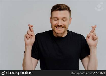 Studio portrait young man with closed eyes making wish, excited guy crossing fingers and asking for good luck while standing against grey studio background. Faith and superstition concept. Young man believer keeping fingers crossed and hoping for best luck