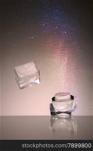 Studio shot of a dropped transparent glass cosmetic jar falling and bouncing off a reflective surface with a fine cloud of colourful cosmetics powder suspended in the air above it