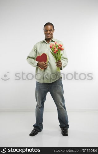 Studio shot of African-American man holding valentine heart and bouquet of tulips smiling and looking at viewer.