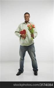 Studio shot of African-American man holding valentine heart and bouquet of tulips smiling and looking at viewer.