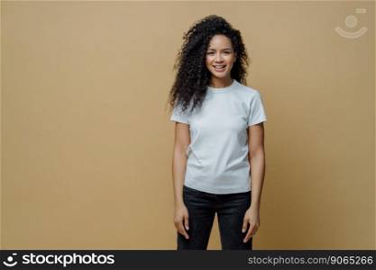 Studio shot of attractive cheerful woman with Afro hairstyle smiles positively, rejoices buying new clothes, wears white t shirt and jeans, enjoys free time for rest, poses against brown background