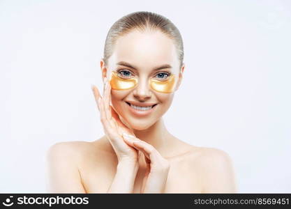 Studio shot of attractive European woman touches face gently, smiles tenderly, applies golden hydrogel patches under eyes, isolated on white background. Eye mask treatment. Anti age procedure