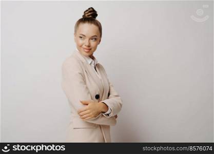 Studio shot of attractive female office manager or business leader in beige suit keeping arms crossed while standing sideways isolated over light background, looking at her co workers with smirk. Office manager in beige suit with crossed arms standing on grey background