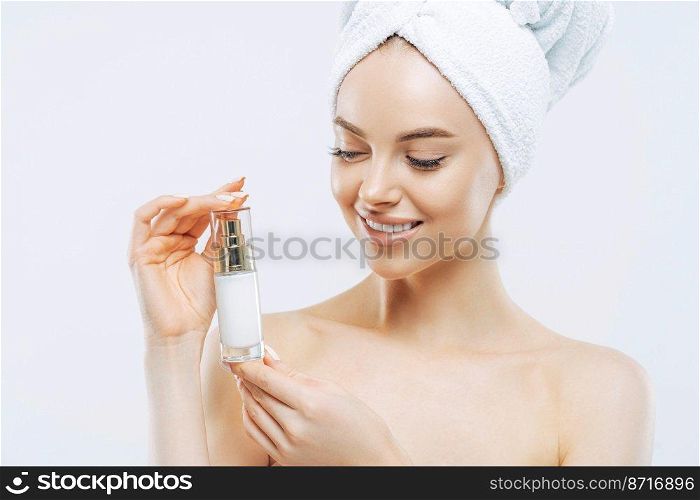 Studio shot of attractive satisfied woman applies moisturizer, holds bottle of skin cream or lotion, wears minimal makeup, poses shirtless against white background, wrapped towel on head. Wellness