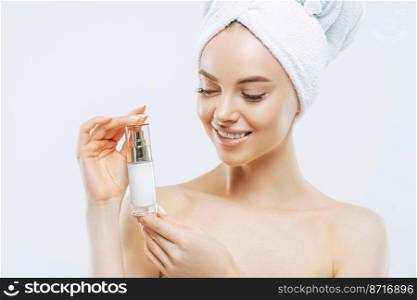 Studio shot of attractive satisfied woman applies moisturizer, holds bottle of skin cream or lotion, wears minimal makeup, poses shirtless against white background, wrapped towel on head. Wellness