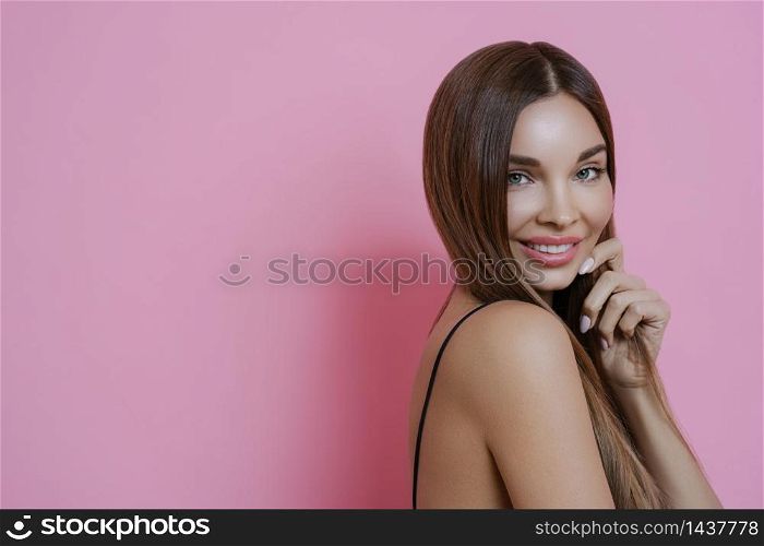 Studio shot of attractive smiling brunette lady, has manicure and dark straight hair, shows bare shoulder, poses against pink background, copy space for your promotional content. Beauty concept