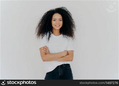 Studio shot of carefree young beautiful woman with Afro hairstyle, keeps arms folded, smiles joyfully, wears casual t shirt and jeans, isolated on white background. People, ethnicity, face expressions