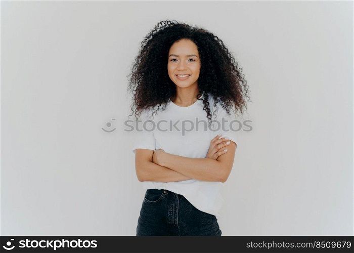 Studio shot of carefree young beautiful woman with Afro hairstyle, keeps arms folded, smiles joyfully, wears casual t shirt and jeans, isolated on white background. People, ethnicity, face expressions
