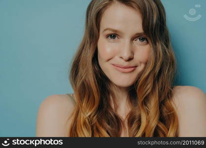 Studio shot of charming shy young woman with wavy brown hair looking at camera, close up portrait timid female with naked shoulders posing isolated over blue background. Women beauty concept. Studio shot of charming shy young woman with wavy brown hair looking at camera with slight smile