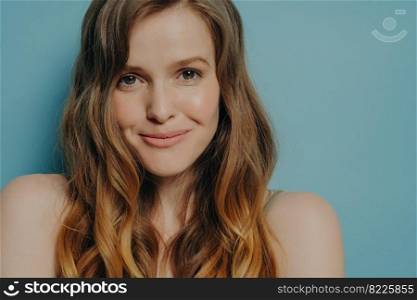 Studio shot of charming shy young woman with wavy brown hair looking at camera, close up portrait timid female with naked shoulders posing isolated over blue background. Women beauty concept. Studio shot of charming shy young woman with wavy brown hair looking at camera with slight smile
