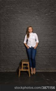 Studio shot of elegant sexy woman in shirt and jeans leaning against black brick wall