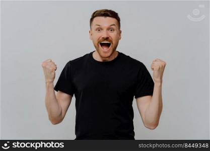 Studio shot of excited happy man in black t shirt reaching personal goals, raising clenched fists and screaming from happiness while celebrating success, standing against grey background. Excited happy man celebrating triumph, reaching personal goals