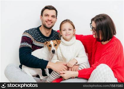 Studio shot of happy family of three family members and dog, embrace and smile happily, have good relationships, isolated on white background. Beautiful small kid with her parents and favourite pet