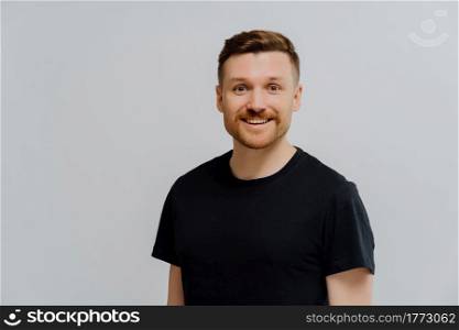 Studio shot of happy handsome ginger man being in good mood, looking at camera with pleasant expression while standing in black tshirt against grey wall. People and positive emotions. Good looking ginger man smiling at camera, expressing positivity