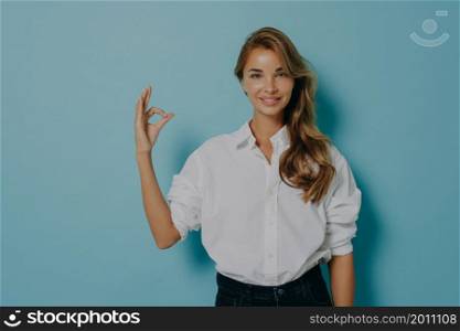 Studio shot of lovely young female model shows okay gesture assures you confirms something is good being confident in quality wears formal white shirt and jeans isolatedover blue background.. Female model shows okay gesture assures you confirms something is good being confident in quality