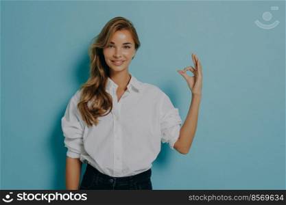 Studio shot of lovely young female model shows okay gesture assures you confirms something is good being confident in quality wears formal white shirt and jeans isolatedover blue background.. Female model shows okay gesture assures you confirms something is good being confident in quality