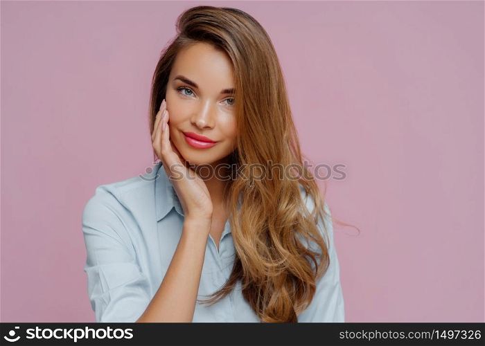 Studio shot of pleasant looking relaxed female model with long hair touches cheek gently, wears elegant shirt, looks straightly at camera, shows her beauty. Pretty businesswoman prepares to work