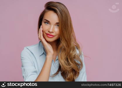 Studio shot of pleasant looking relaxed female model with long hair touches cheek gently, wears elegant shirt, looks straightly at camera, shows her beauty. Pretty businesswoman prepares to work