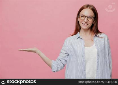 Studio shot of pleased Caucasian woman keeps palm raised, wears optical glasses, demonstrates something over copy space, holds blank space, has pleasant smile on face, poses indoor over pink wall